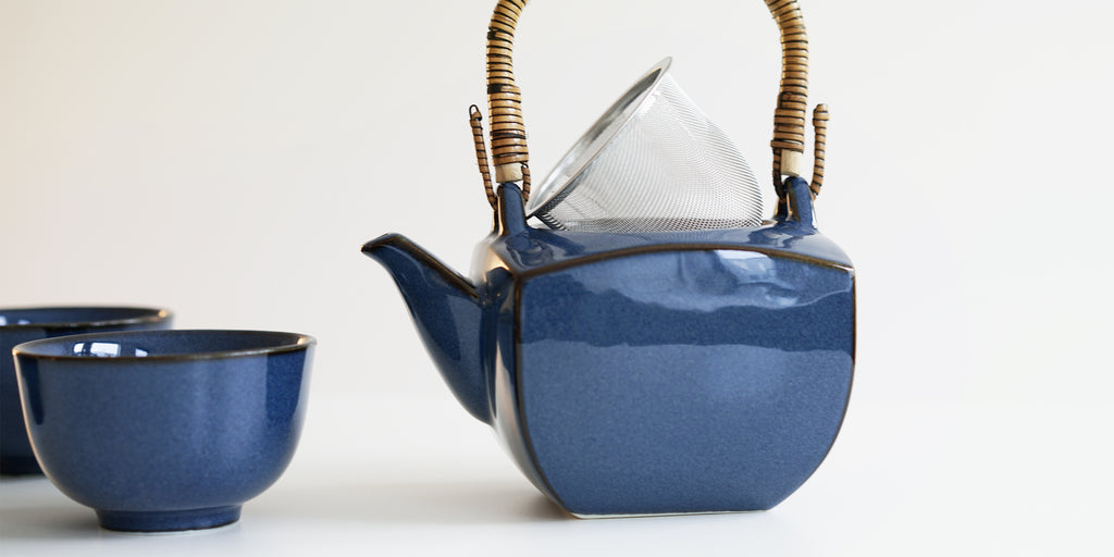 Side view of a dark blue ceramic tea pot. The lid is off and the strainer is set asked on top of the pot.