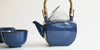 Side view of a dark blue ceramic tea pot. The lid is off and the strainer is set asked on top of the pot.