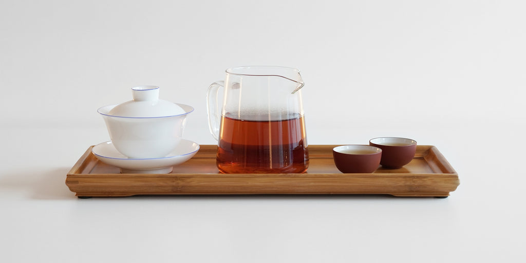 From left to right, a gaiwan, a glass server full of tea, and two teacups, all placed on a bamboo tea tray.