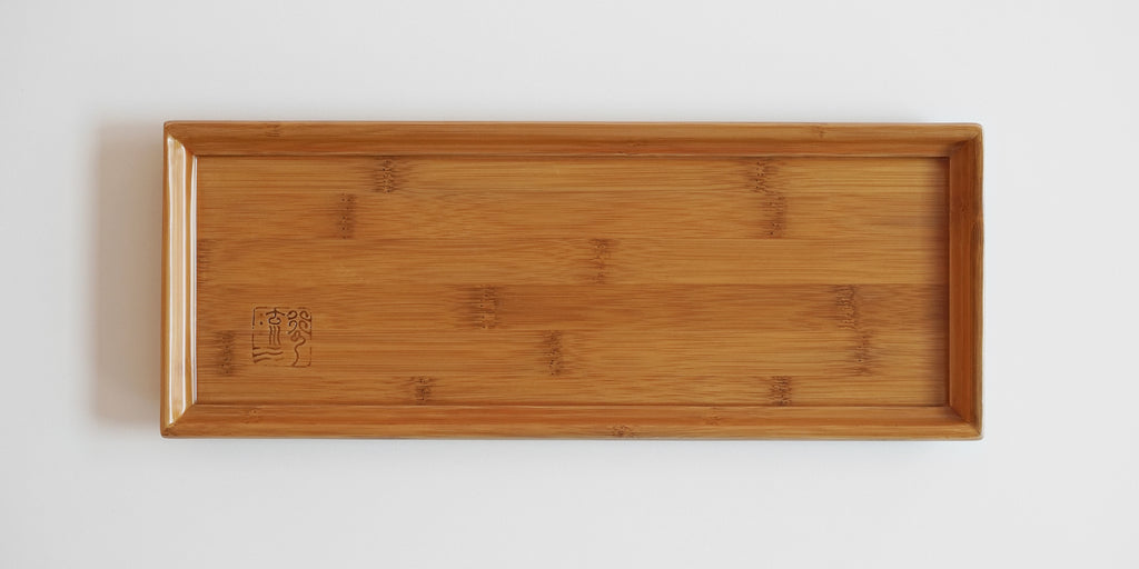 Top view of a bamboo tea tray. A lip runs around the outside to prevent spillage, and a maker's mark is in the bottom left corner.