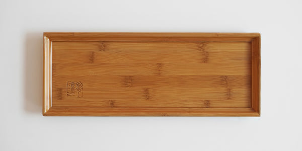 Top view of a bamboo tea tray. A lip runs around the outside to prevent spillage, and a maker's mark is in the bottom left corner.