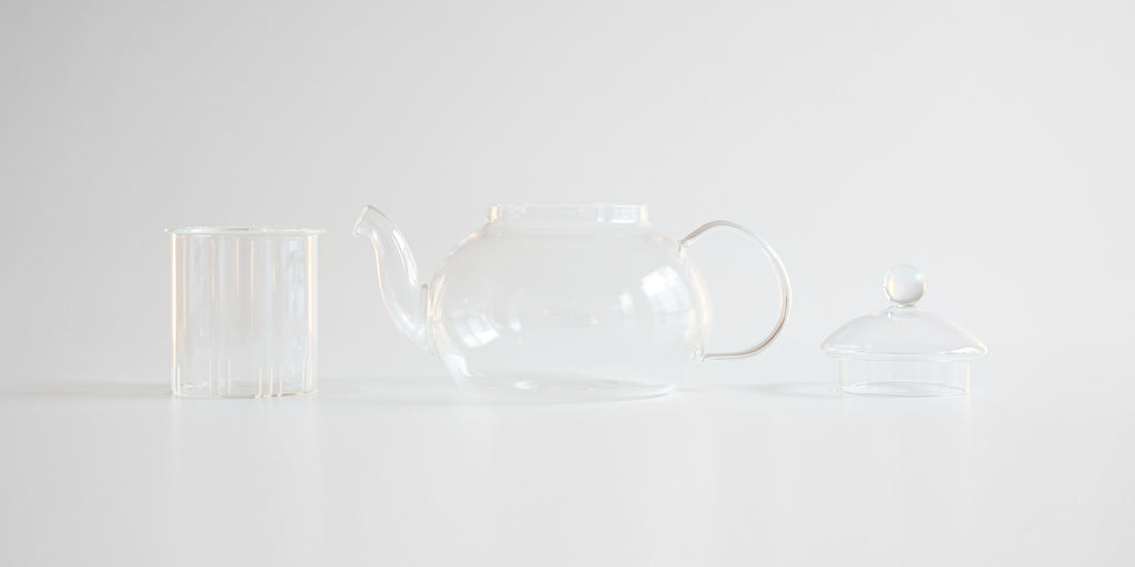 Side view of each part of a glass teapot laid out. From left to right, the infuser, the pot, and the lid.