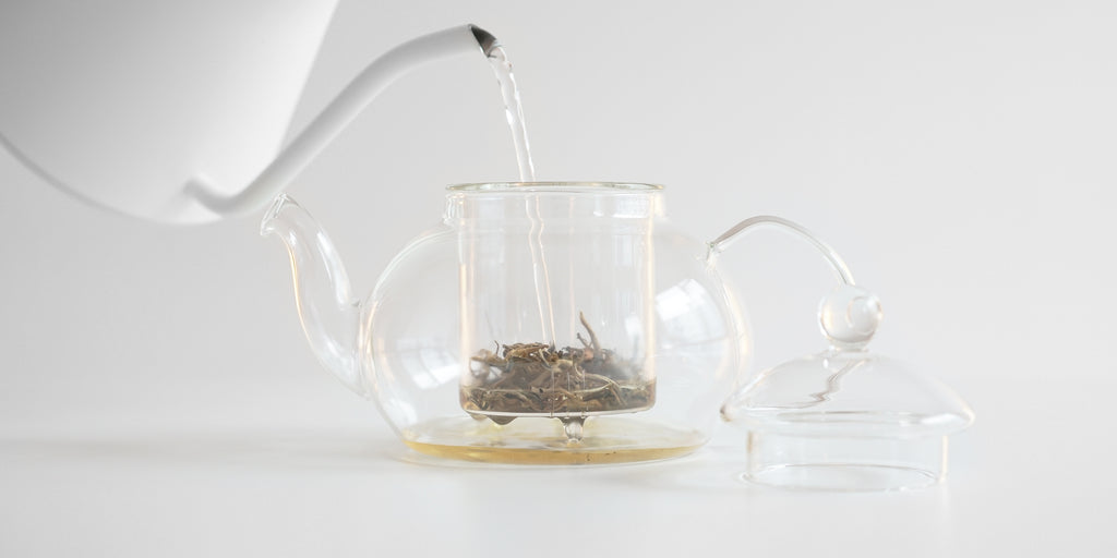 Side view of water being added to a glass teapot, with leaves in the nested glass infuser.