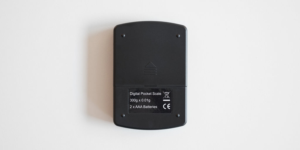 Bottom of a matte black scale. It has a sliding panel to insert batteries into, and four small feet on the corners.