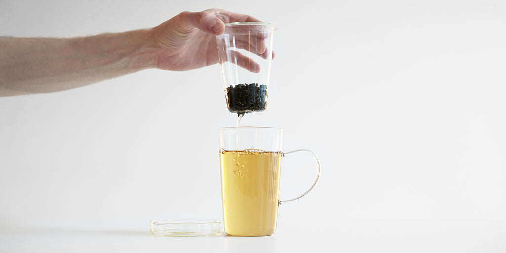Side view of a glass cup filled with tea. A hand is removing the glass strainer, allowing the remaining tea to flow out of the slits in the infuser.