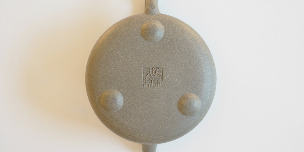 The bottom of a gray duanni yixing teapot, with three small feet around the base, and two maker's marks stamped into it, one in the middle of the base, and the other at the bottom of the handle.