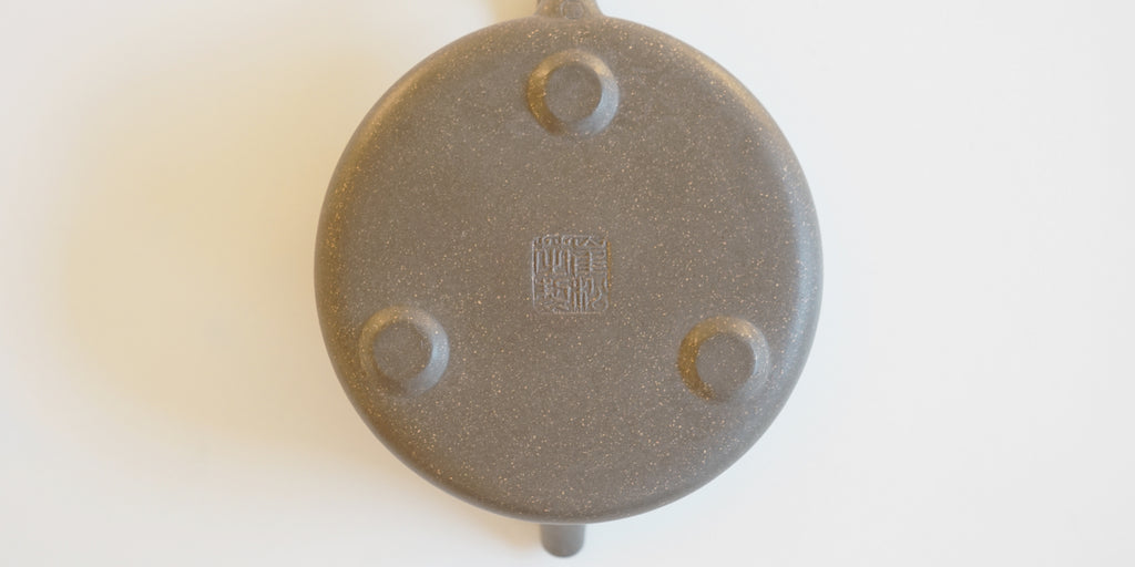 The base of a gray duanni yixing pot. It has three small feet around the outer edge, and a maker's mark stamped into the middle.