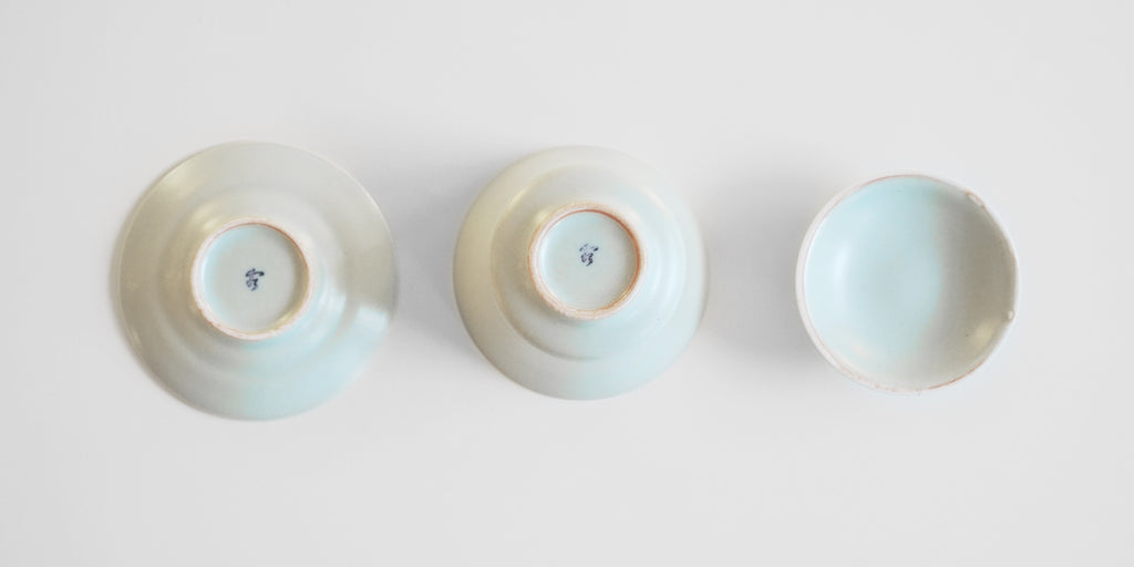 Top down view of the undersides of each piece of a blue-green celadon gaiwan. The bowl and plate each have a maker's mark painted into the foot.
