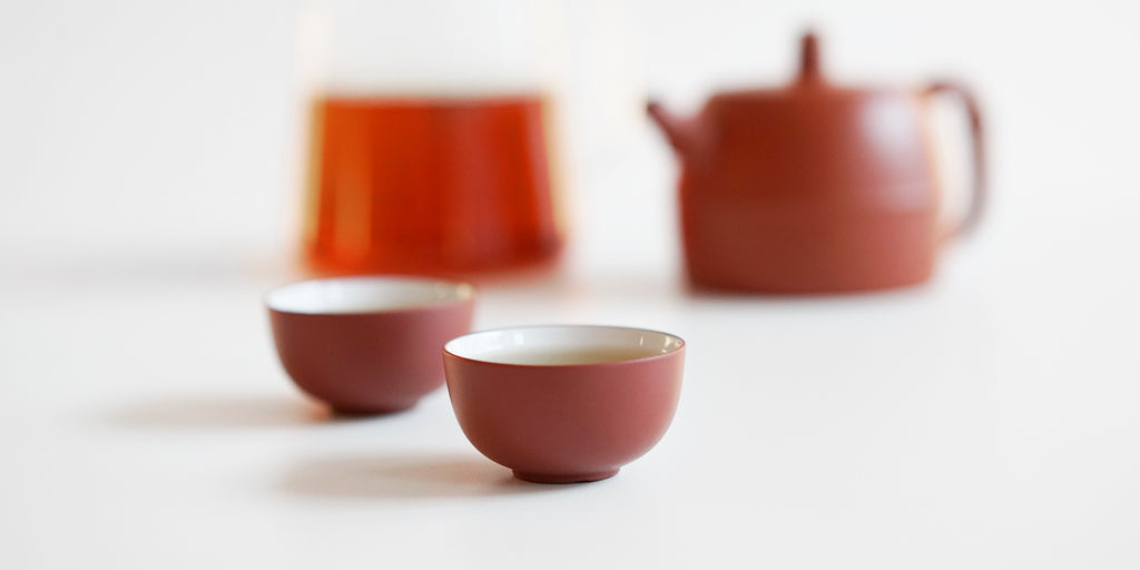 Two teacups, terracotta-colored on the outside, white on the inside. A teapot and a pitcher full of tea are out of focus in the background.