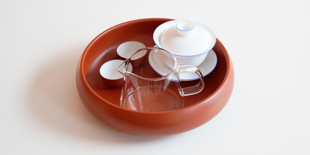 A white gaiwan, a glass server, and two teacups in a round terracotta-colored tea tray.