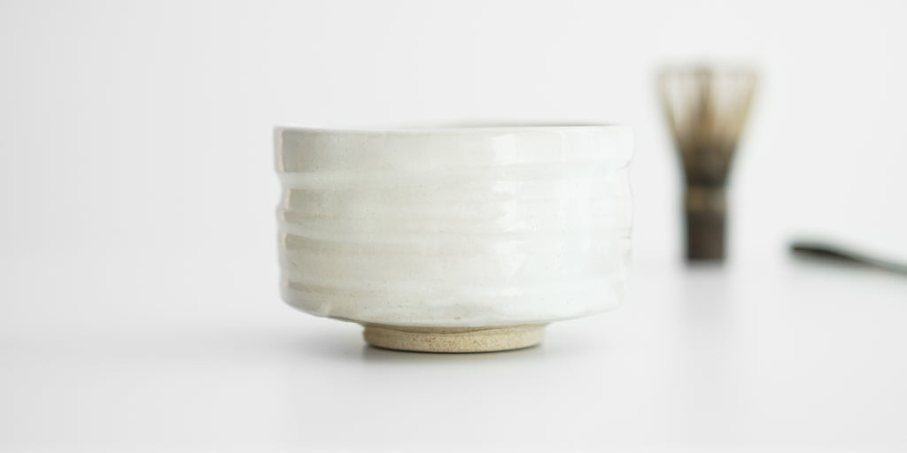 Side view of a white bowl with an unglazed foot. A matcha whisk and scoop are in the background.