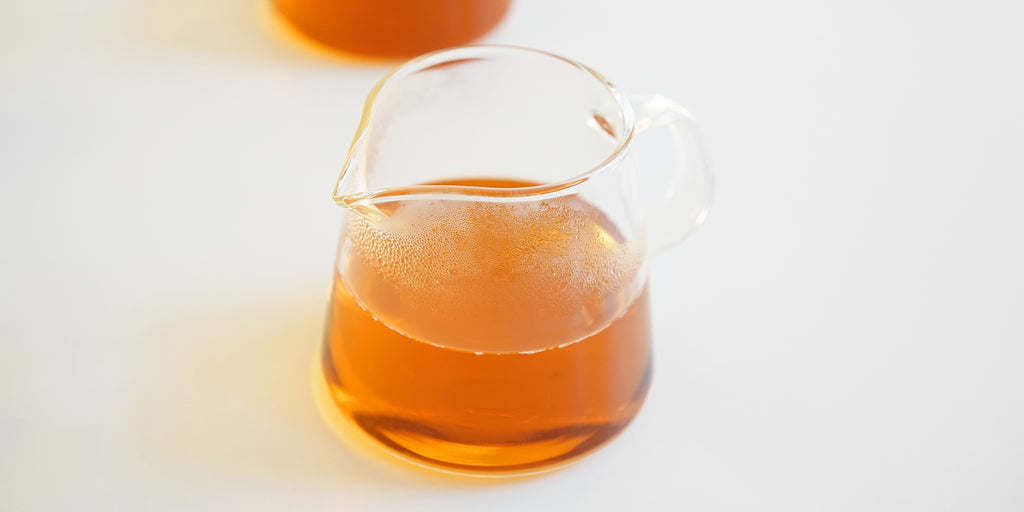 A glass server with tea in it, steam gathering above the tea. A second server is blurred in the background.