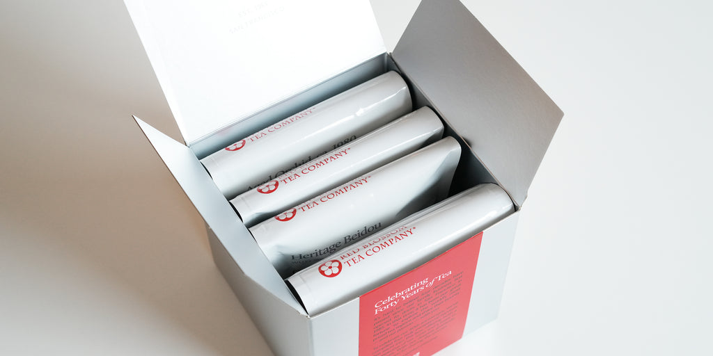 A silver box with 4 white bags of tea showing the Red Blossom Tea Company logo inside.