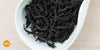 A white dish containing dry black tea leaves. An orange circle in the bottom left corner reads "MX"