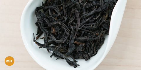 A white dish containing dry black tea leaves. An orange circle in the bottom left corner reads "MX"