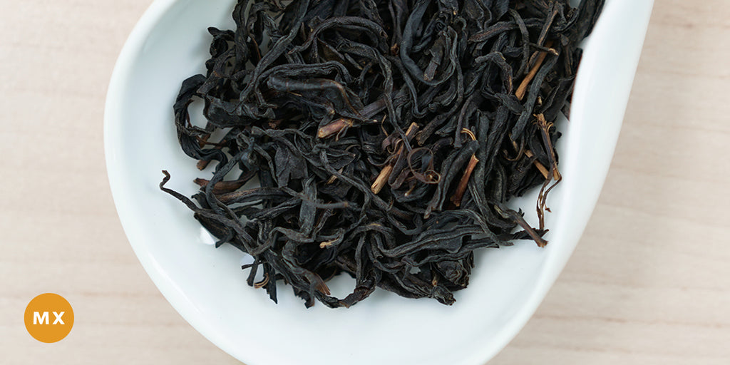 A white dish containing dry black tea leaves. An orange circle in the bottom left corner reads "MX"