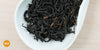 A white dish containing dry black tea leaves. An orange circle in the bottom left corner reads "MX"
