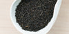 A white dish containing dry black tea leaves.