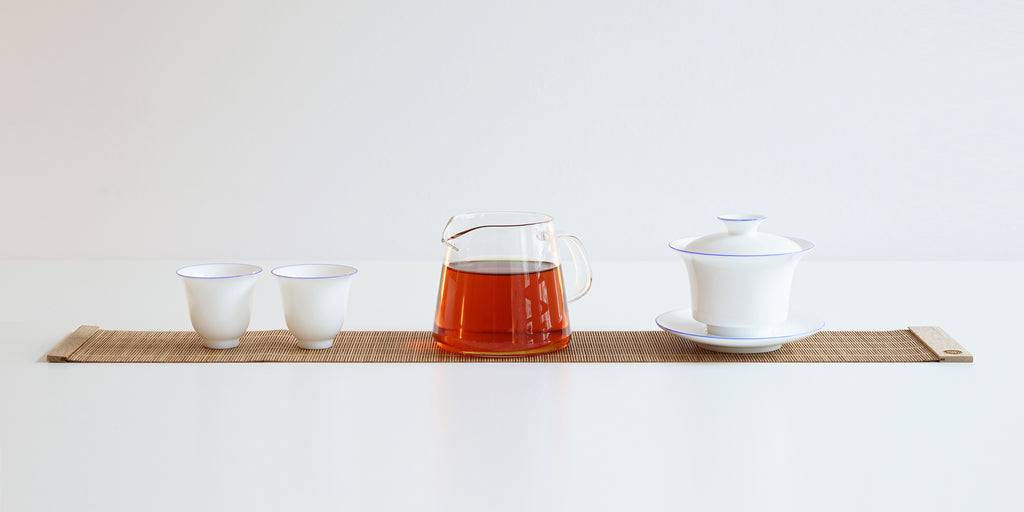 From left to right, two white teacups, a glass server full of tea, and a gaiwan all sit on top of a roll-up bamboo table runner.