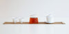 From left to right, two white teacups, a glass server full of tea, and a gaiwan all sit on top of a roll-up bamboo table runner.