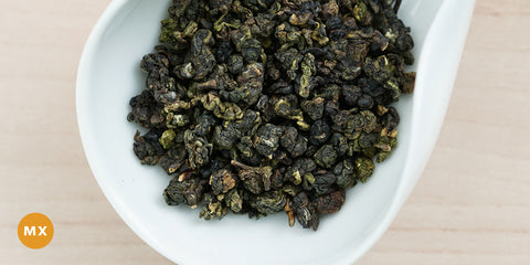 A white dish containing dry Formosa oolong tea leaves. An orange circle in the bottom left corner reads "MX"