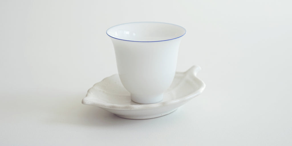 A white teacup with a blue rim set on top of a white trivet in the shape of a leaf.