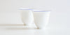 Two white teacups, curving up at the base and out towards the thin lip, with a blue ring painted around the edge.