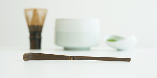 Close up of a purple bamboo matcha scoop, with a whisk, white bowl, and dish full of matcha out of focus in the background.