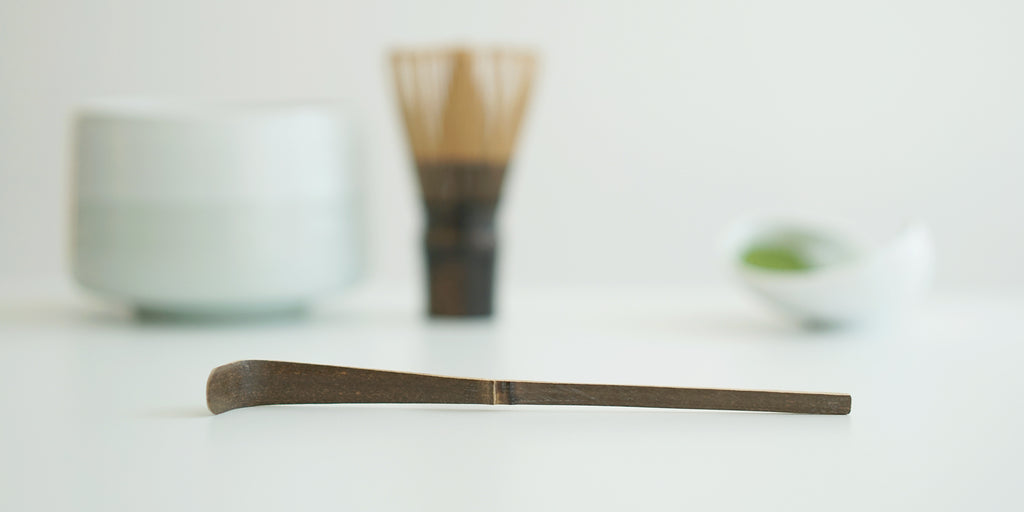 A dark brown colored matcha scoop made of purple bamboo resting on a white surface. A straight-sided white bowl, matching matcha whisk, and a white dish of matcha are in the background.