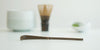 A dark brown colored matcha scoop made of purple bamboo resting on a white surface. A straight-sided white bowl, matching matcha whisk, and a white dish of matcha are in the background.