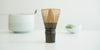 A matcha whisk resting on a white surface. A small back string is tied around the base of the prongs. A white bowl with a matcha scoop resting on top of it and a white dish of matcha are in the background.