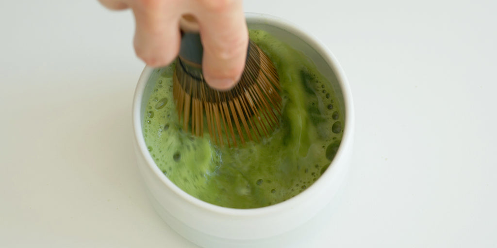 A hand uses a purple bamboo matcha whisk to froth a bowl of matcha.