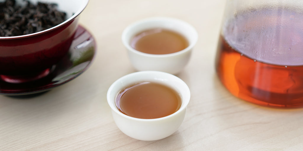 Two small teacups of aged Tieguanyin tea, with a gaiwan in the background on the left and a glass server on the right.