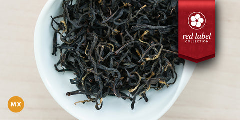 A white dish containing dry black tea leaves. A red ribbon in the top right corner reads “Red Label Collection”, and an orange circle in the bottom left corner reads "MX"