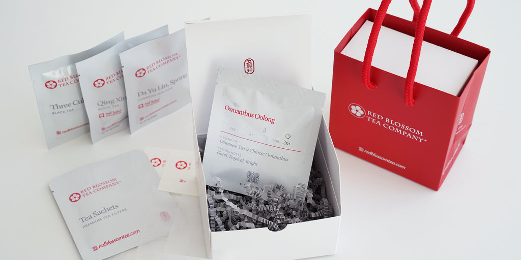 A white gift box containing 4 sample packs of tea and a pack of tea sachets to brew them with. On the right, another box is inside a small branded red bag.
