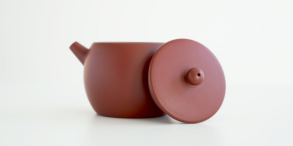Side view of a terracotta-colored yixing teapot. The lid is facing the camera, resting against the handle.