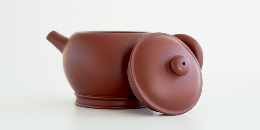 Side view of a terracotta-colored yixing teapot. The lid is facing the camera, resting against the handle.