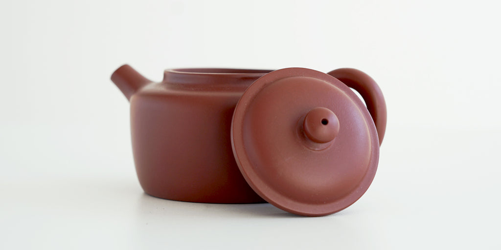 Side view of a terracotta-colored yixing teapot. The lid is facing the camera, resting against the handle.