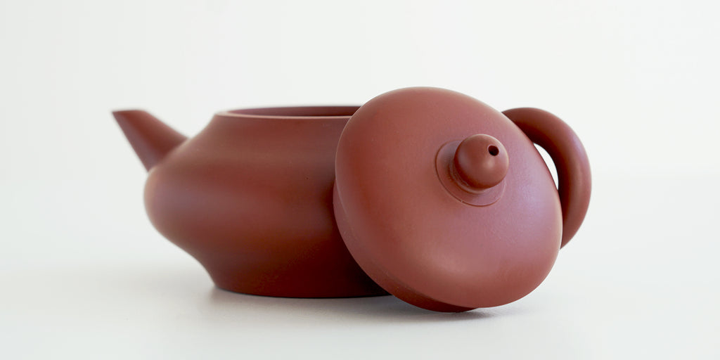 Side view of a terracotta-colored yixing teapot. The lid is facing the camera, resting against the handle.