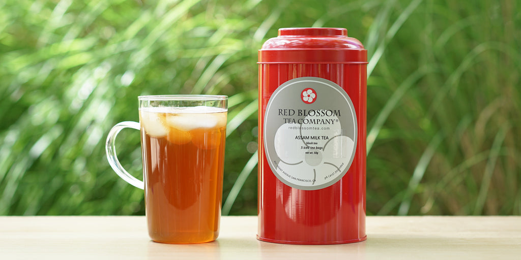 A red airtight canister of Assam Milk Tea next to a glass of brewed tea, with greenery in the background.
