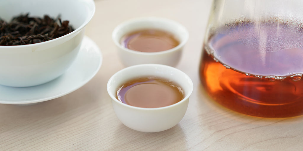 Two small teacups of black tea, with a gaiwan in the background on the left and a glass server on the right.