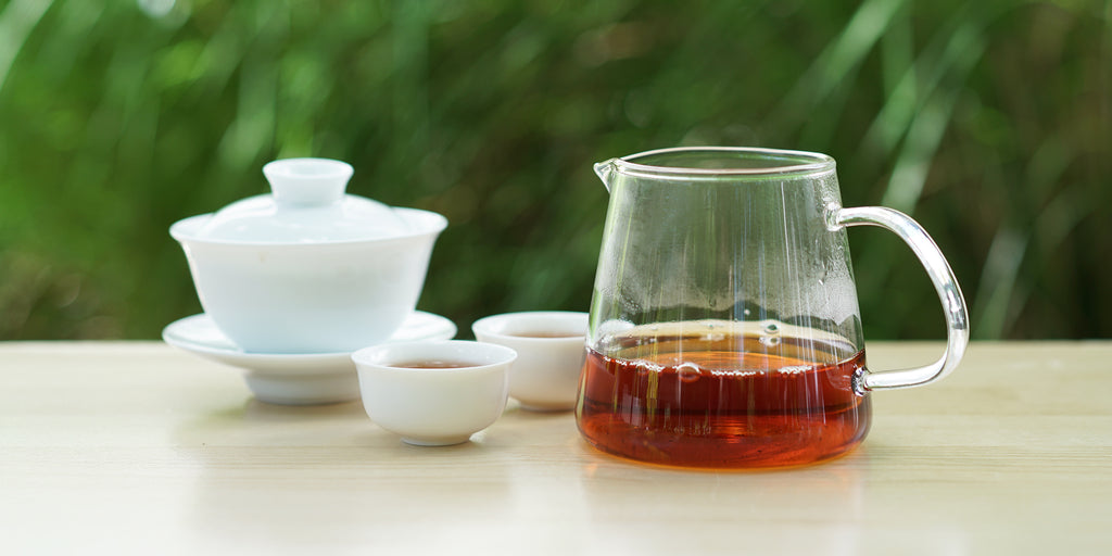 From left to right, a gaiwan, two teacups, and a glass server. The teacups and server have black tea in them.