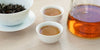 Two small teacups of black tea, with a gaiwan in the background on the left and a glass server on the right.