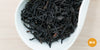 A white dish containing dry black tea leaves. An orange circle in the bottom right corner reads "MX"