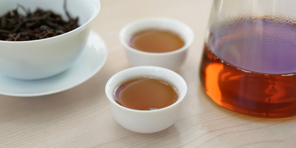 Two small teacups of black tea, with a gaiwan in the background on the left and a glass server on the right.