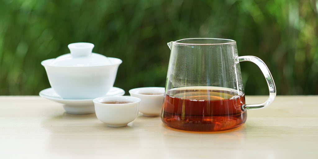 From left to right, a gaiwan, two teacups, and a glass server. The teacups and server have black tea in them.