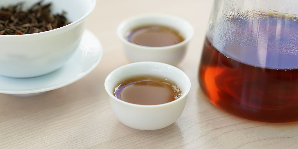 Two small teacups of black tea, with a gaiwan in the background on the left and a glass server on the right.