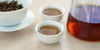Two small teacups of black tea, with a gaiwan in the background on the left and a glass server on the right.