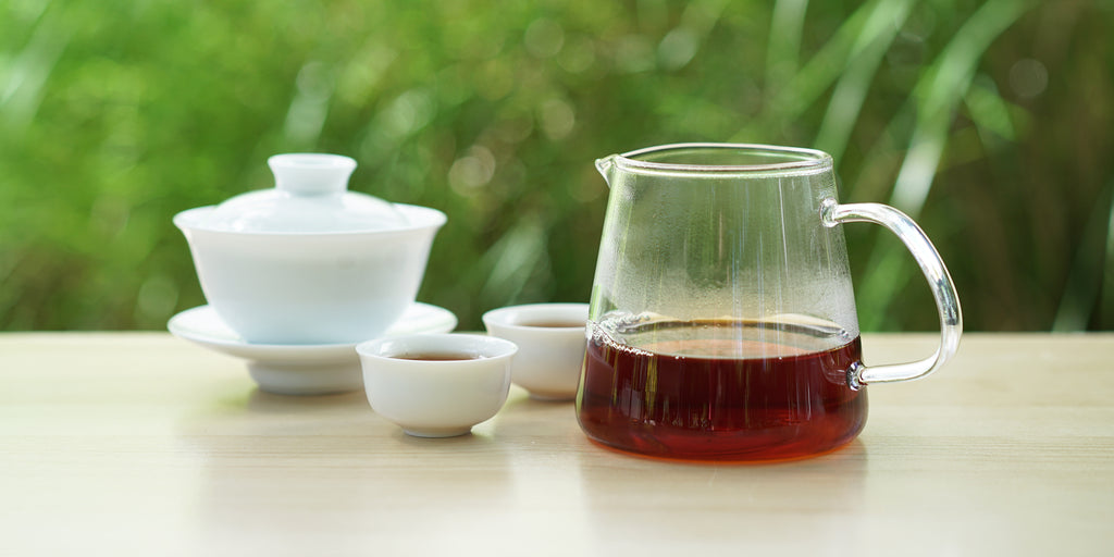 From left to right, a gaiwan, two teacups, and a glass server. The teacups and server have black tea in them.