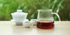 From left to right, a gaiwan, two teacups, and a glass server. The teacups and server have black tea in them.